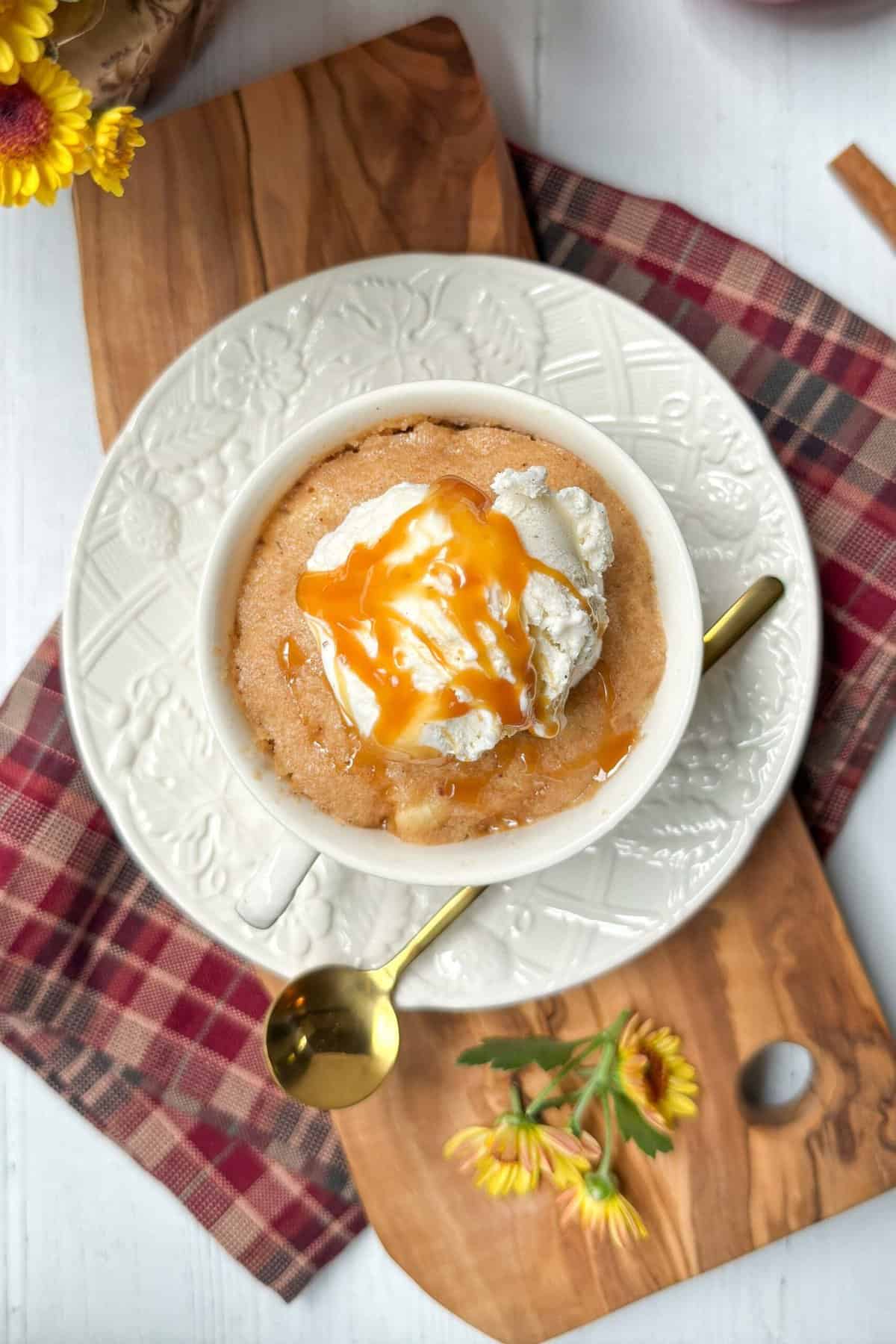 A top-down view of a mug cake in a white mug topped with a scoop of vanilla ice cream and drizzled with caramel sauce. The mug is on a decorative white plate with a golden spoon beside it. The plate sits on a wooden cutting board with a checked cloth and yellow flowers nearby.