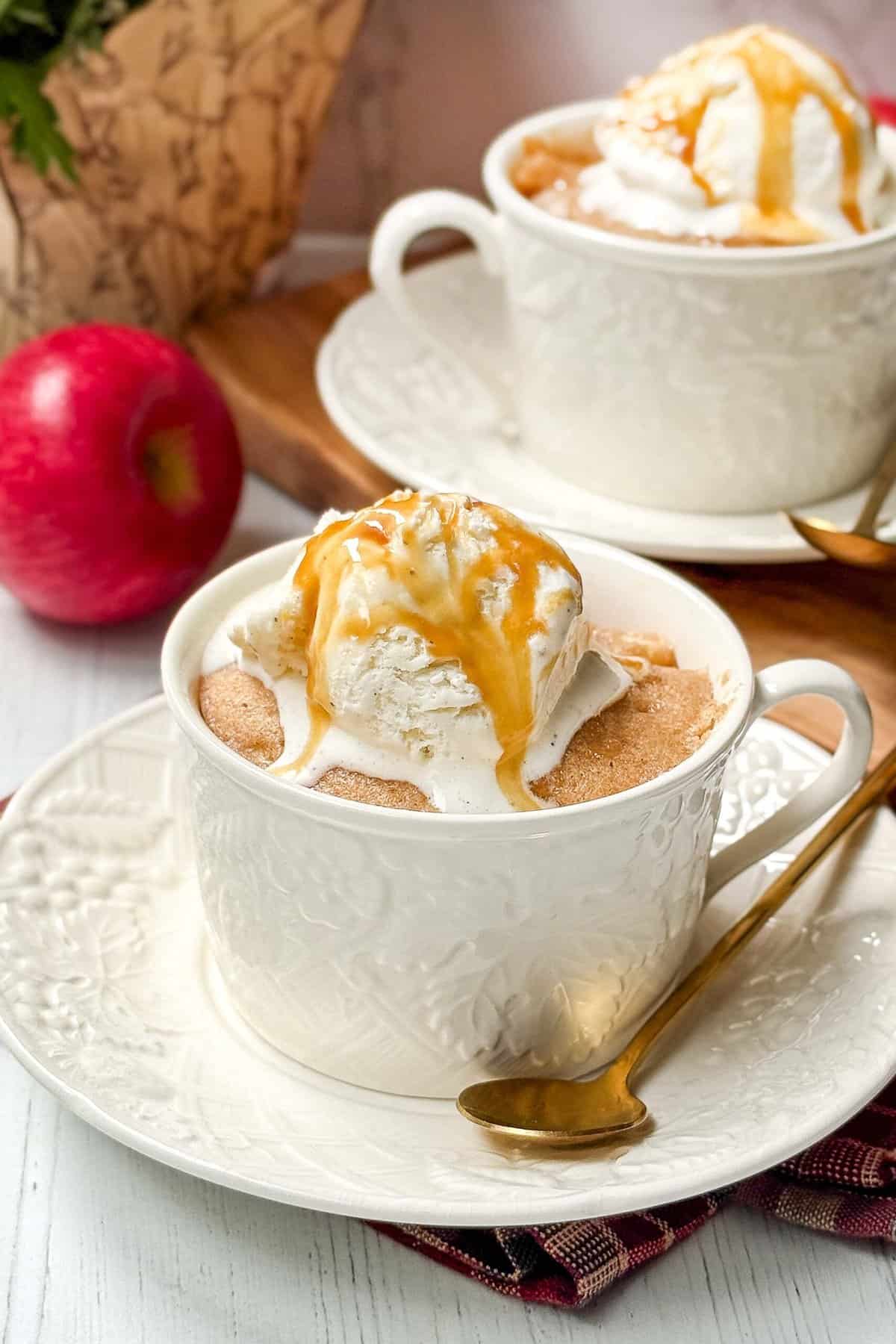 Two apple mug cakes presented in white mugs sit atop matching saucers. Each cake is topped with a scoop of vanilla ice cream and drizzled with caramel sauce. A shiny gold spoon accompanies the setting next to an apple and a wooden cutting board in the background.