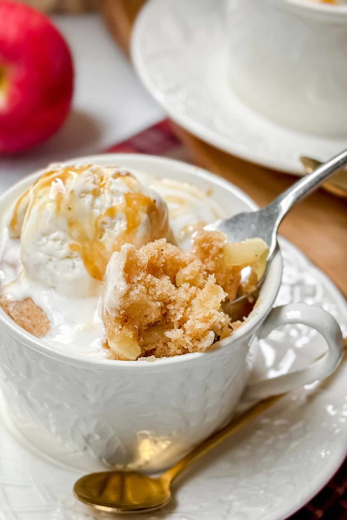 A close-up of a white teacup filled with a mug cake topped with a scoop of vanilla ice cream and drizzled with caramel sauce. A spoon is taking a bite from the delightful apple mug cake. A red apple and another teacup are visible in the background.