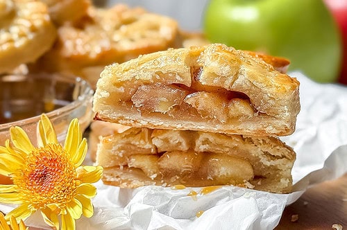 A close-up of two stacked apple pie slices on a wooden board, reminiscent of classic apple hand pies, surrounded by yellow flowers and fresh apples. The pie crust is golden and flaky, revealing a juicy apple filling.