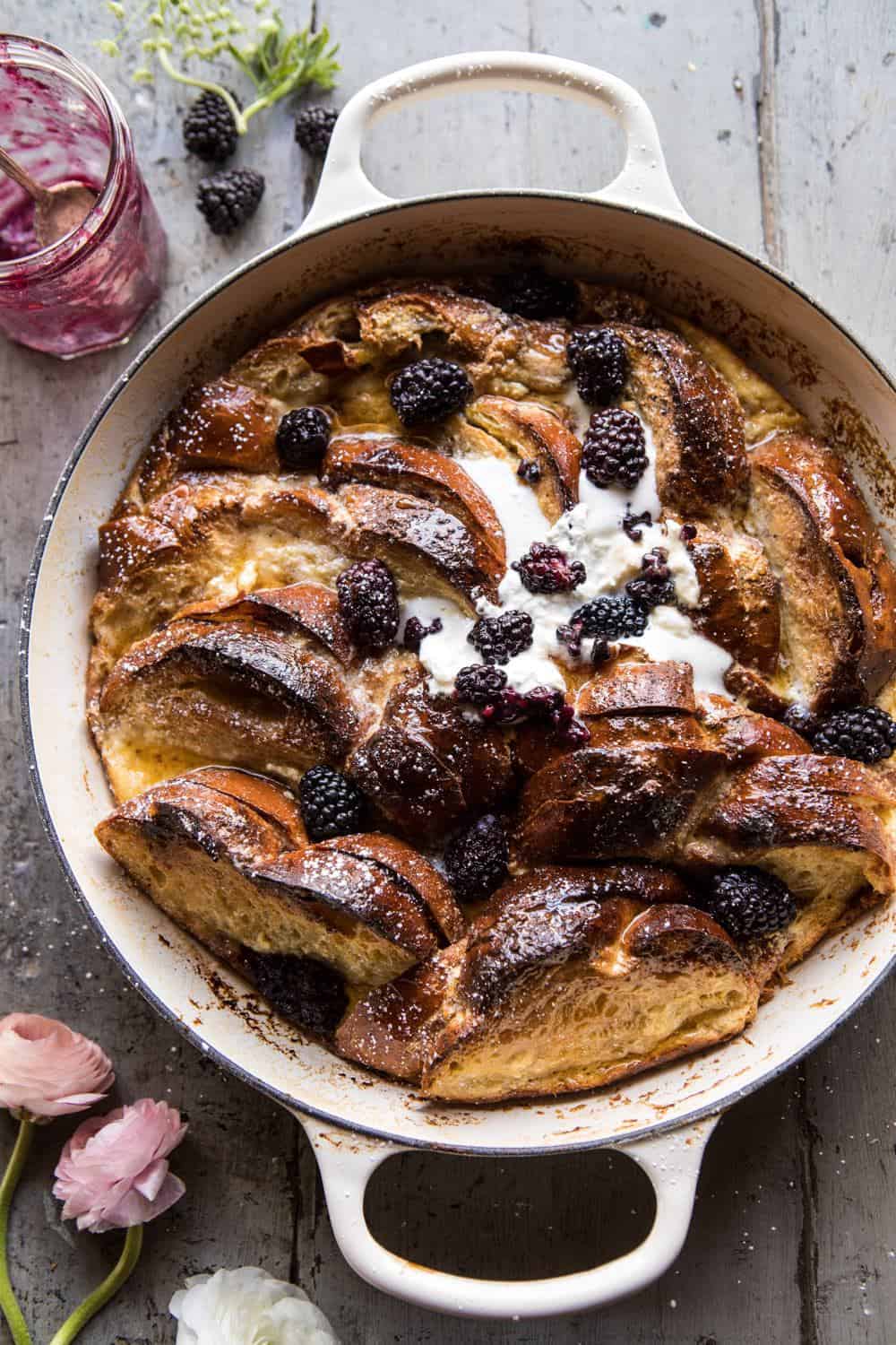 A baked bread pudding in a round dish, topped with fresh blackberries and a dollop of cream. The rustic wooden background is decorated with fresh flowers and a jar of blackberry preserve.