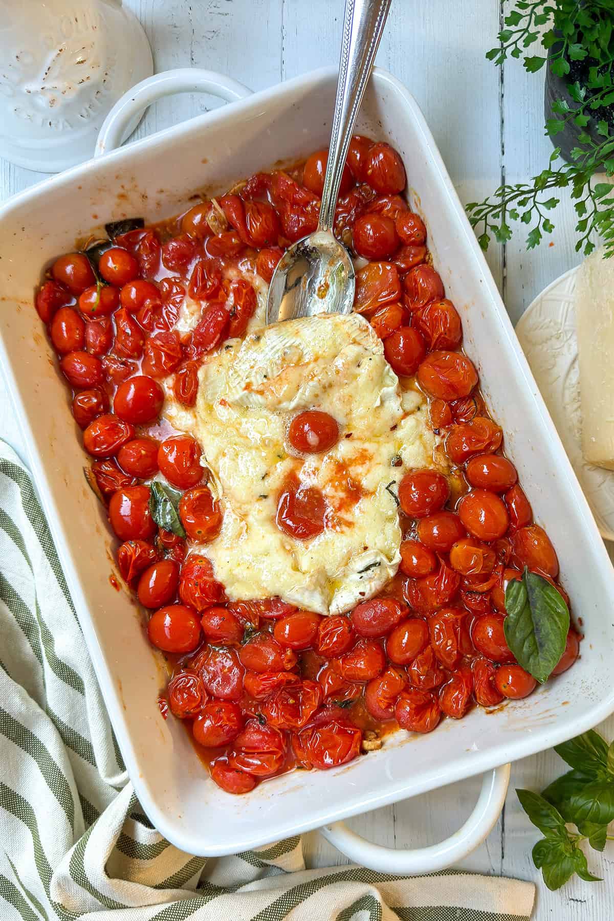 A white baking dish filled with roasted cherry tomatoes and a block of melted brie cheese garnished with fresh basil leaves, with a serving spoon on top.