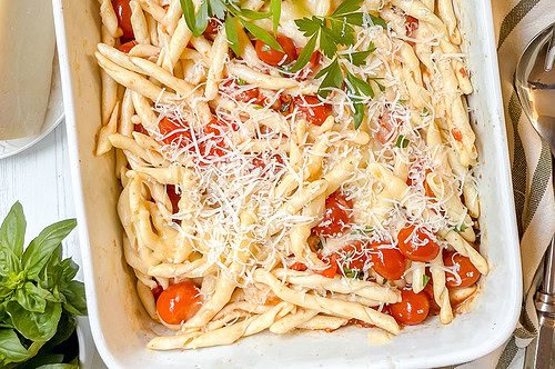 A white baking dish filled with twisted pasta mixed with cherry tomatoes, topped with grated cheese and a sprig of parsley. Fresh herbs and a striped cloth linen are nearby on a white table.