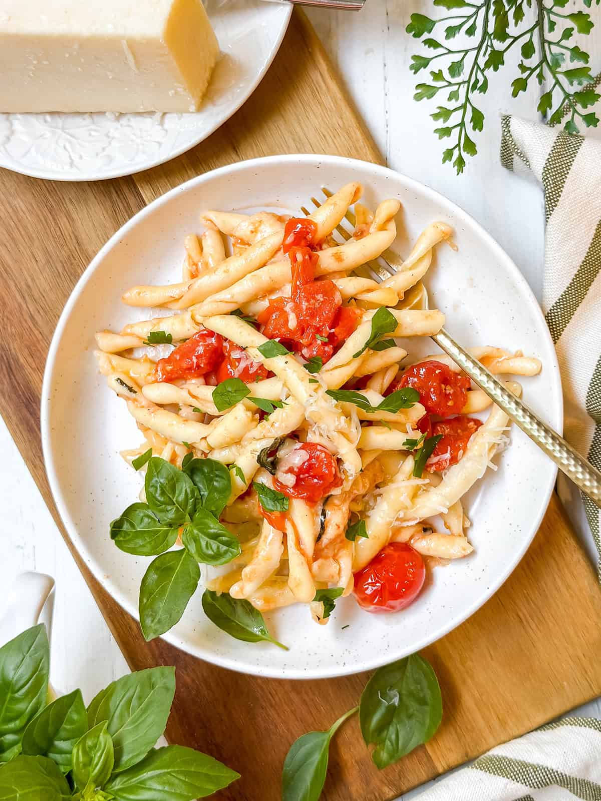 A plate of pasta with baked brie tomato sauce, cherry tomatoes, basil, and grated cheese sits on a wooden board alongside fresh herbs and a wedge of cheese.