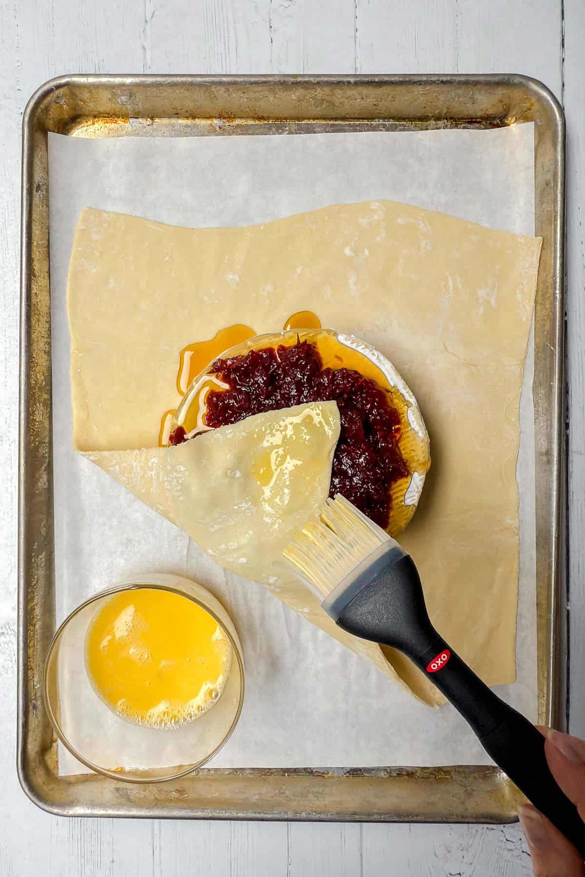 Folding bottom left corner of puff pastry over honey cranberry topped Brie wheel and brushing egg wash over the top.