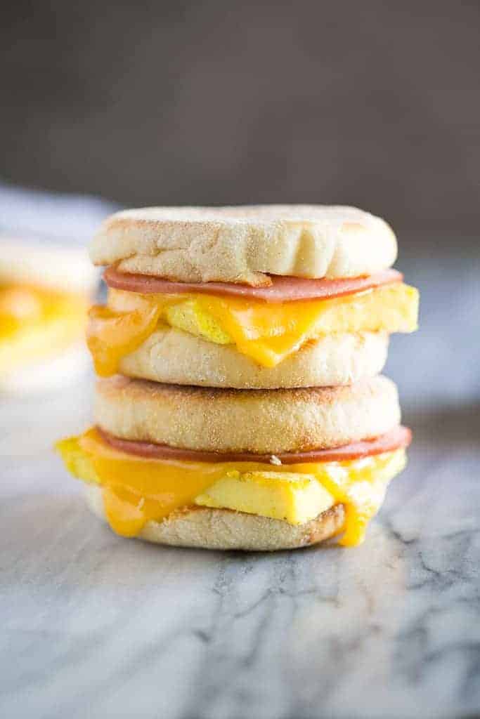 A stack of three breakfast sandwiches with English muffins, melted cheese, egg, and ham on a marble surface.