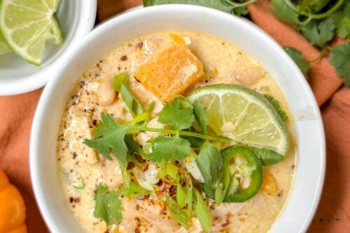A bowl of butternut chicken chili garnished with lime slices, cilantro, and red pepper flakes. The creamy soup is set on a table with decorative pumpkins, an orange cloth, and a side bowl with lime slices. Text overlay reads "Easy Butternut Chicken Chili Recipe - Irresistibly Delicious!