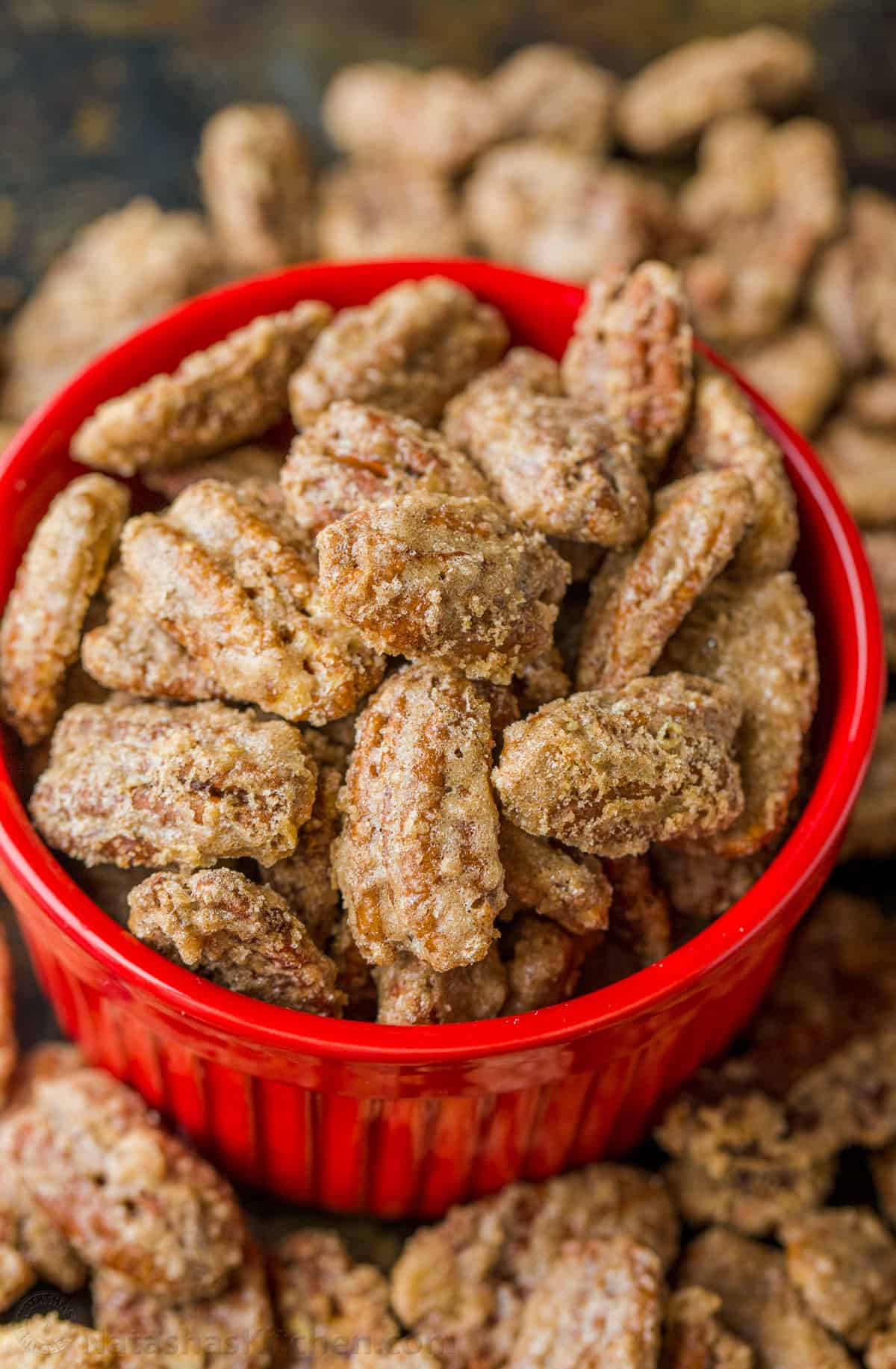A close-up of a red bowl filled with sugar-coated pecans, surrounded by more sugared pecans scattered around. The nuts appear crunchy and glistening, suggesting a sweet, glazed texture.