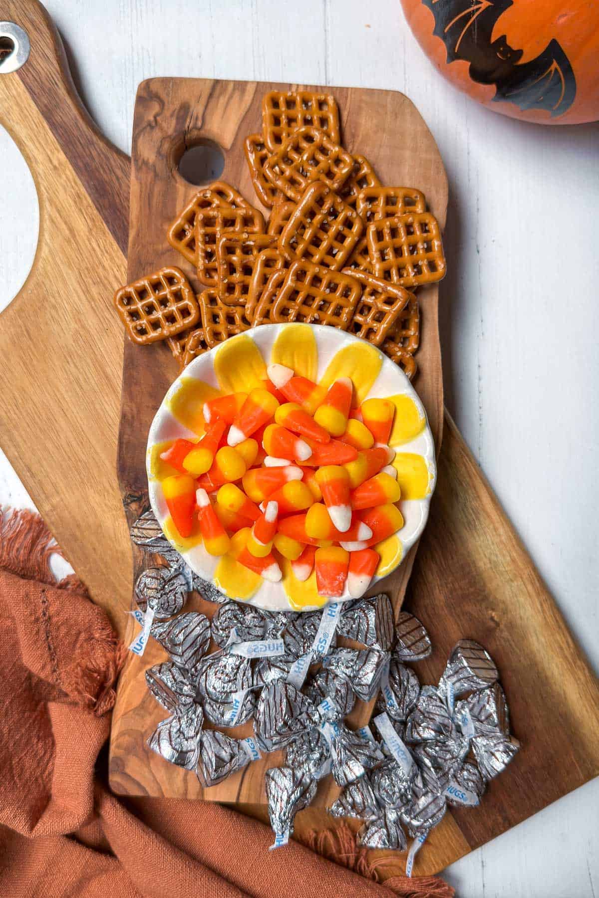 A wooden board with a bowl of candy corn surrounded by square pretzels and foil-wrapped chocolates. A carved pumpkin and an orange cloth are nearby, creating a festive, autumnal setting.
