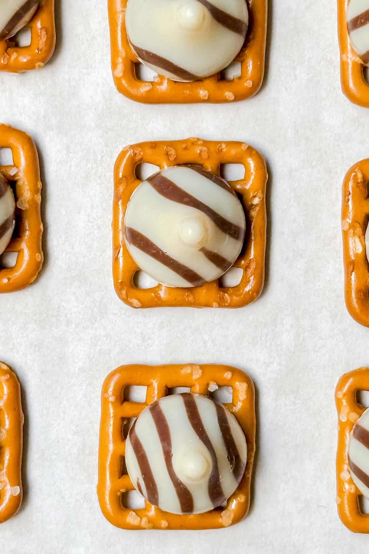 Close-up of several pretzels topped with round striped chocolate candies on a white background. The pretzels are square-shaped with a light brown color, and each has a white and dark brown striped chocolate piece atop.