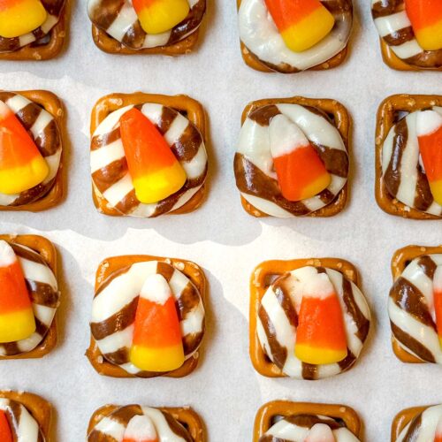 Square pretzels topped with striped chocolate and a candy corn each are arranged in rows on a baking sheet. The pretzels have a glossy appearance, and the overall look is festive and colorful.