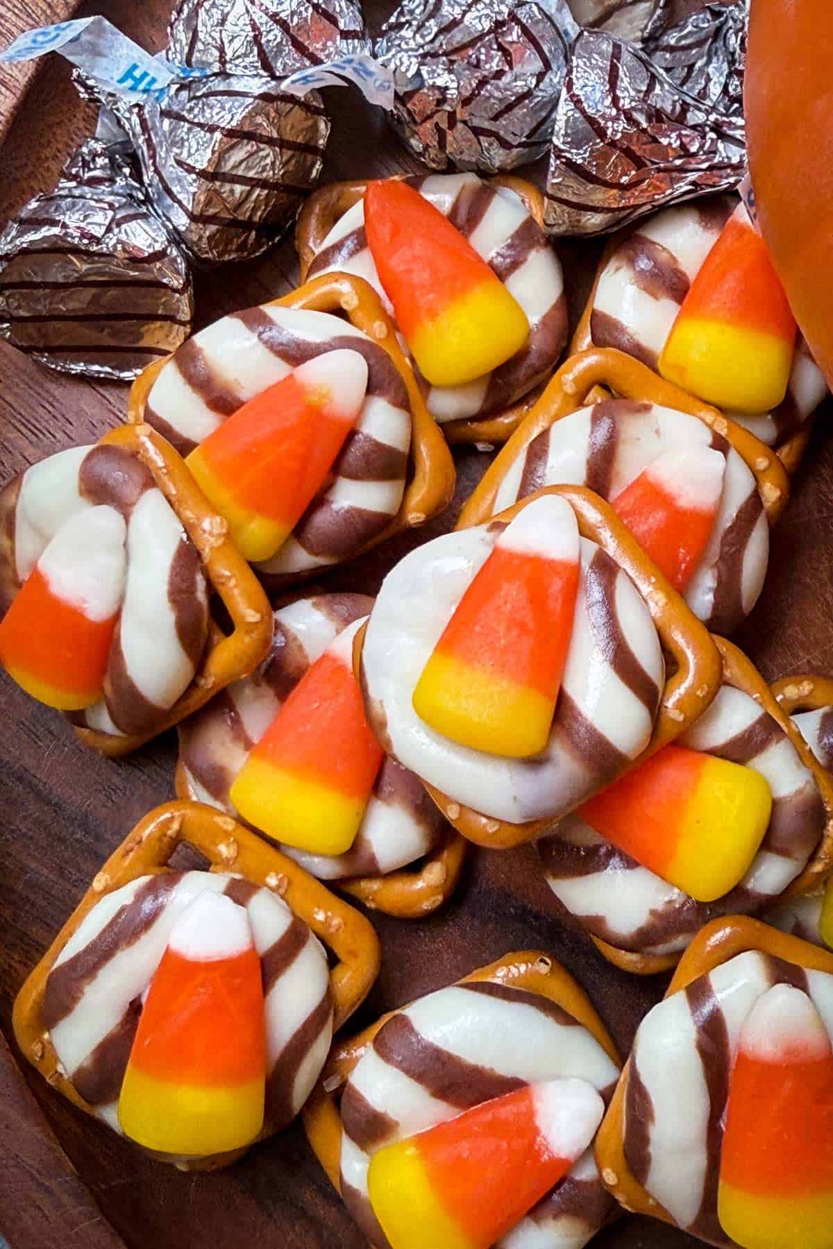 A wooden tray with pretzels topped with striped chocolate kisses and candy corn. Silver foil wrappers are scattered in the background.