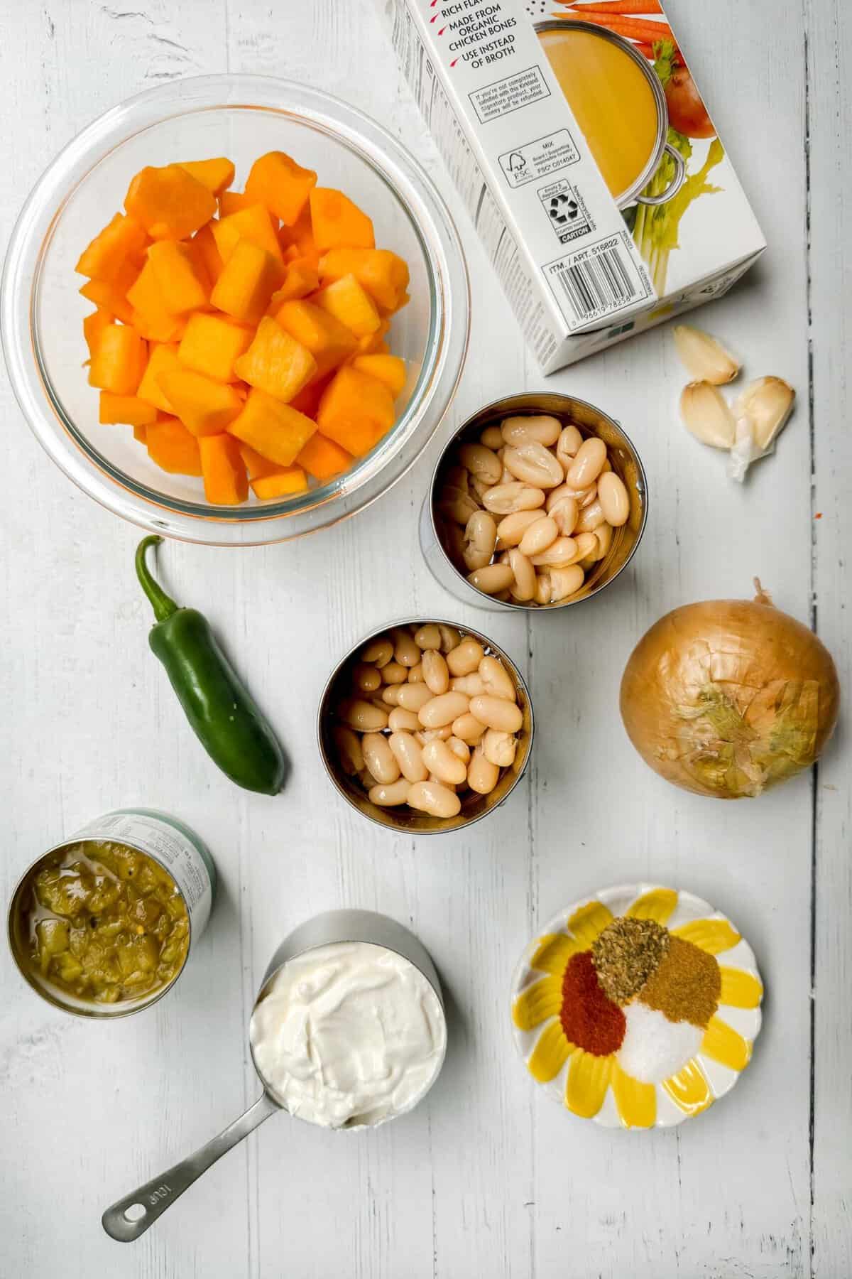 Top-down view of ingredients for a chicken chili on a white wooden surface. Items include a bowl of chopped butternut squash, a carton of chicken broth, canned white beans, garlic cloves, an onion, a jalapeño, a can of green chilies, a bowl of spices, and a measuring cup of sour cream.