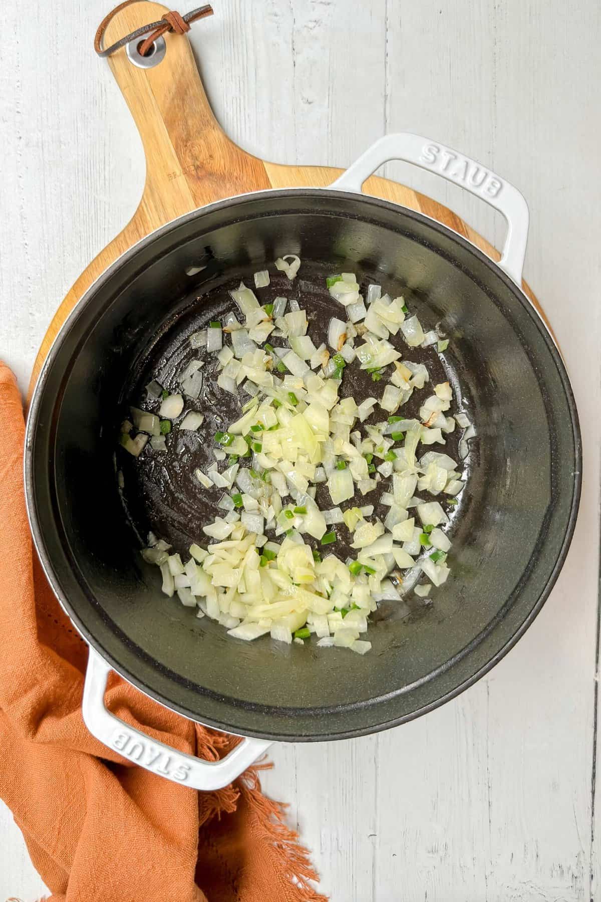 A white cast iron pot on a wooden board, containing chopped onions and jalapeños being sautéed. An orange cloth is placed next to the pot on a light wooden surface.