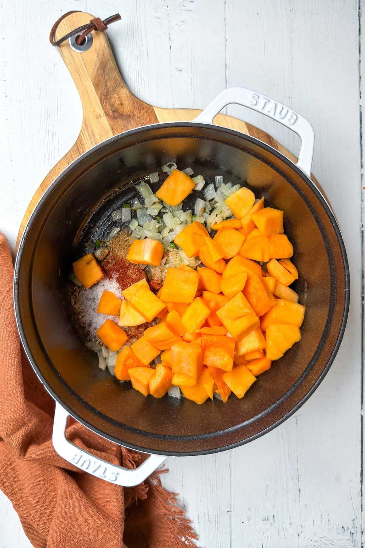 A top-down view of a pot with diced butternut squash, chopped onions, and seasonings, placed on a round wooden cutting board with a brown cloth napkin beside it. The pot is white with "STAUB" written on the handles.