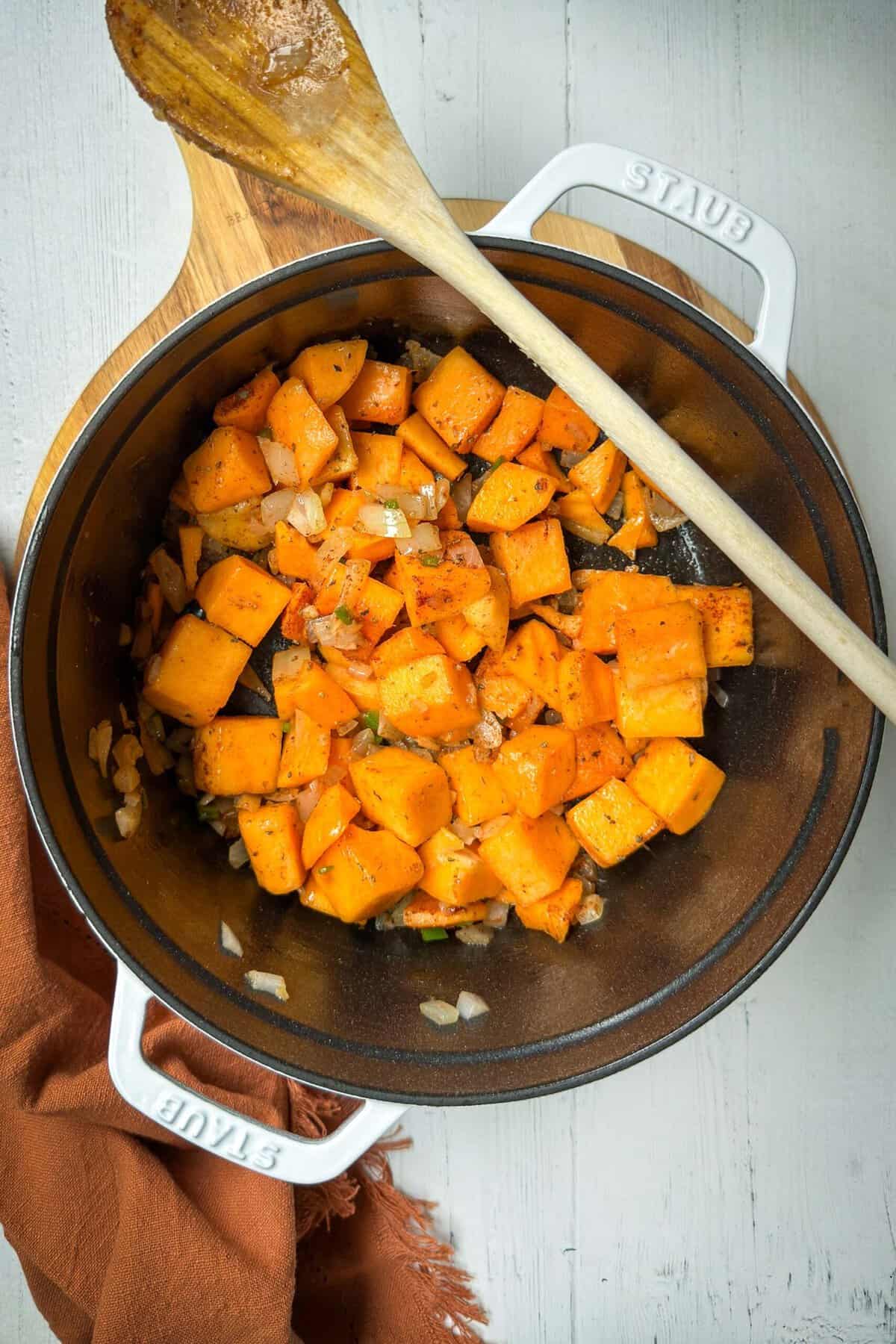 A white Staub pot on a wooden board contains diced butternut squash with onions and spices. A wooden spoon rests across the top of the pot. An orange napkin is partially visible to the left side of the pot. The background is a light-colored surface.