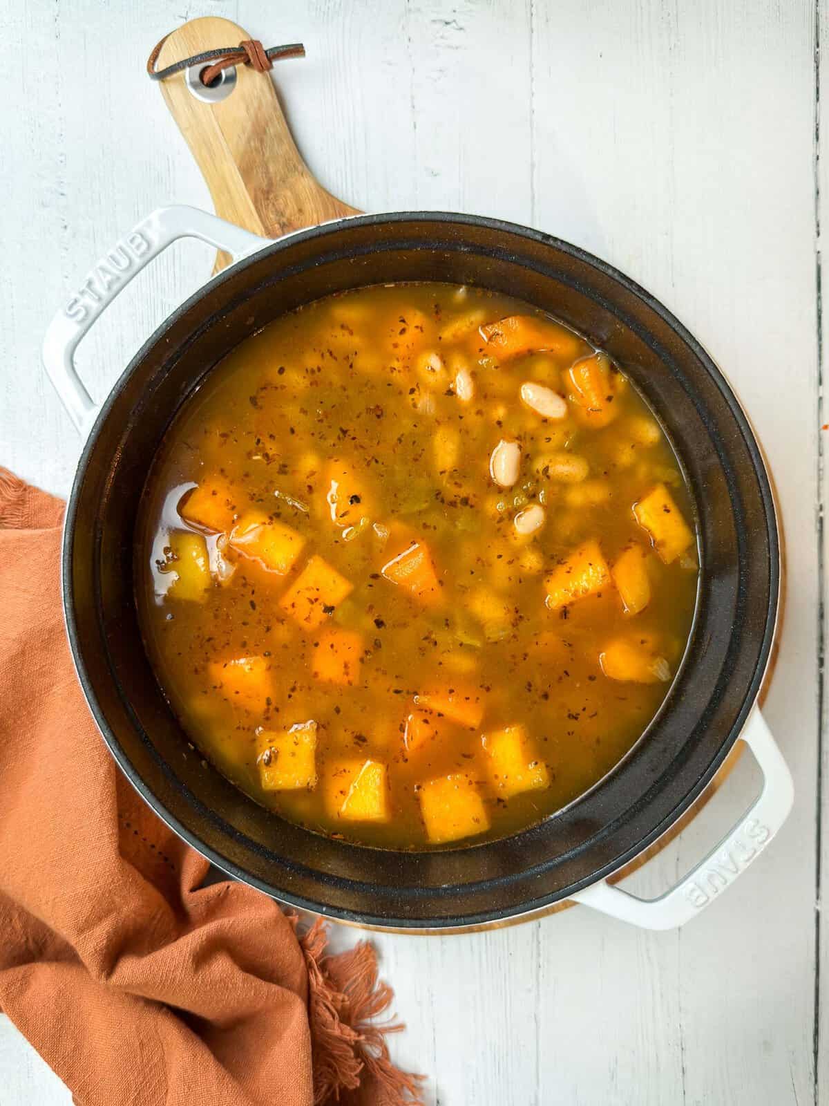 A pot of chili filled with chunks of orange squash, white beans, and spices, sits on a white wooden surface. The pot is cast iron with white handles, and a rust-orange cloth with fringed edges lies beside it.