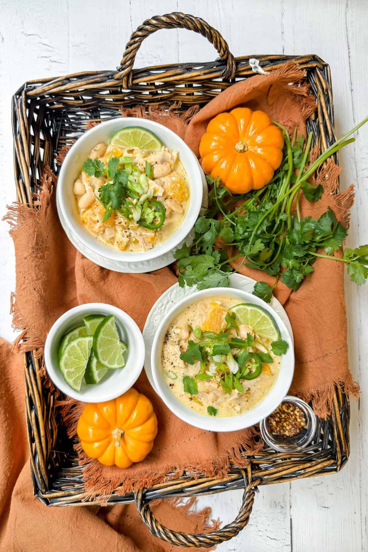 A wicker tray holds two bowls of creamy white chicken chili, garnished with lime, cilantro, and jalapeños. Fresh cilantro, lime slices, two small pumpkins, and a bowl of crushed red pepper flakes accompany the bowls on the tray, which is set over a rustic white wooden surface.