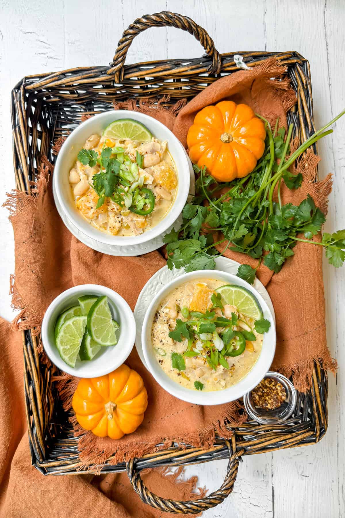 Two bowls of butternut white chicken chili with lime, cilantro, and pumpkin garnish in a rustic fall basket