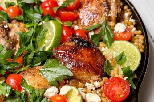 A skillet brimming with succulent chicken thighs, cherry tomatoes, lime wedges, feta cheese, and parsley crowns a bed of fluffy couscous. A bowl with a plate peeks into view on a white surface.