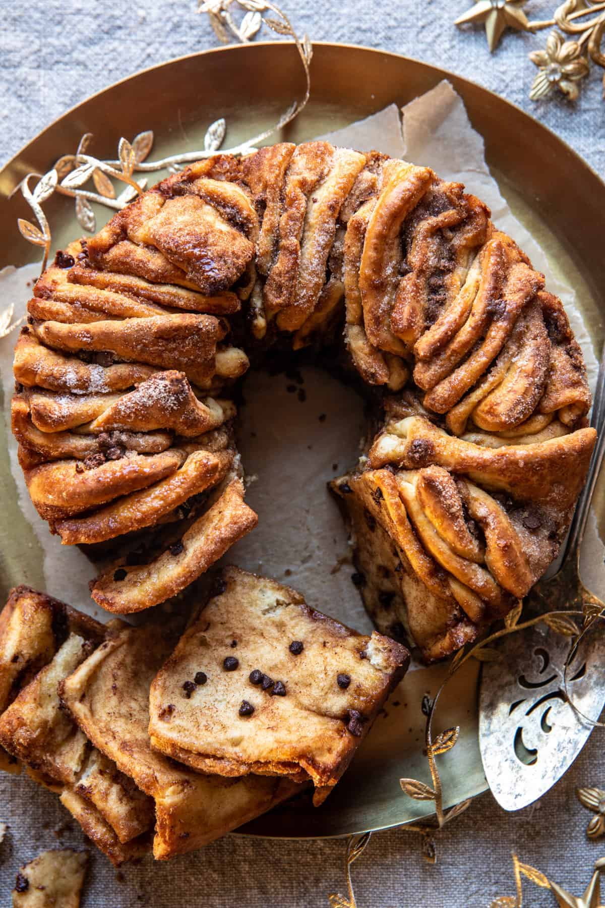 A cinnamon pull-apart bread topped with chocolate chips is served on a decorative round tray. A section is sliced, revealing its layered texture. The bread is placed on parchment paper, with a decorative serving utensil nearby.