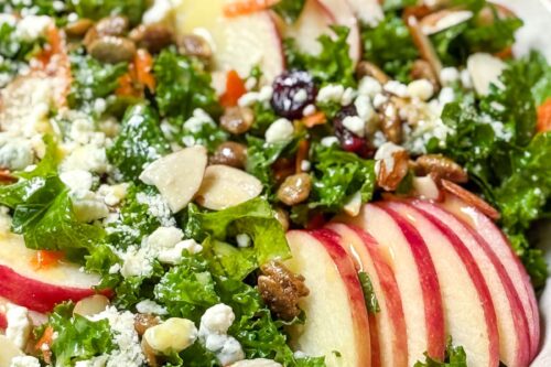 A vibrant kale salad in a bowl features fresh leaves, slices of red apple, crumbled cheese, sliced almonds, and various seeds. A jar of yellow dressing is visible in the background, adding a fresh touch to the scene.