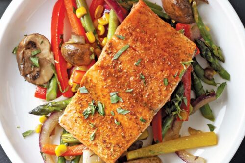 A plate of seasoned salmon atop a colorful mix of vegetables, including red and yellow bell peppers, asparagus, mushrooms, red onions, and corn, garnished with fresh herbs.