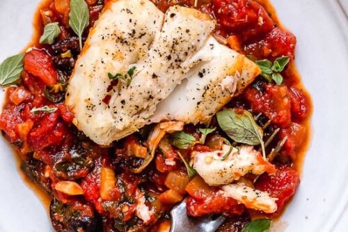 A plate of baked white fish on a bed of tomato-based sauce with black olives and fresh herbs. A fork rests on the plate, and a striped cloth is positioned next to it.