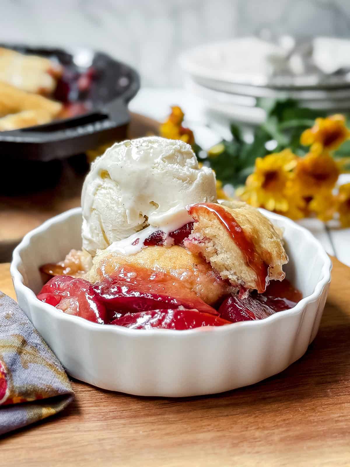 A serving of plum cobbler topped with vanilla ice cream with the skillet dessert in the background.