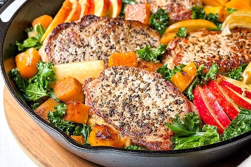 A cast-iron skillet filled with seasoned pork chops, sautéed kale, sliced apples, and cubed squash. In the background, a decorative white oil dispenser and a bowl of red and green apples on a white wooden surface.