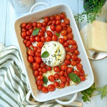 White baking dish with roasted cherry tomatoes, fresh basil leaves, and a round of baked brie cheese in the center.