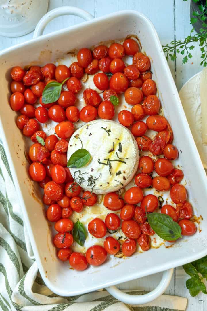 White baking dish with roasted cherry tomatoes, fresh basil leaves, and a round of baked brie cheese in the center.