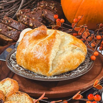 Golden baked brie in puff pastry on a serving board with pumpkin bread in the background.