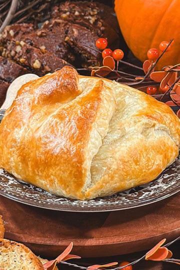 Golden baked brie in puff pastry on a serving board with pumpkin bread in the background.