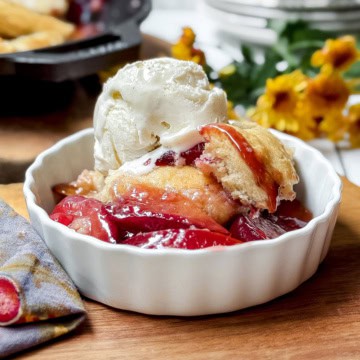 Closeup of plum cobbler serving topped with vanilla ice cream with the skillet dessert in the background.