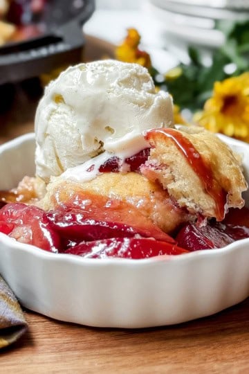 Closeup of plum cobbler serving topped with vanilla ice cream with the skillet dessert in the background.