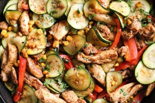A skillet filled with a colorful stir-fry featuring sliced zucchini, red bell peppers, corn, and seasoned strips of chicken. The ingredients are garnished with fresh herbs, creating a vibrant and appetizing dish on a blue wooden surface.