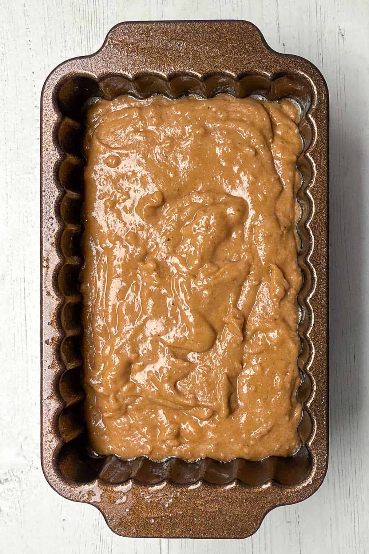 A rectangular baking pan filled with light brown sweet potato bread batter, ready to be baked. The batter has a smooth texture and is spread evenly in the pan, which features a scalloped edge. The pan rests on a white wooden surface.