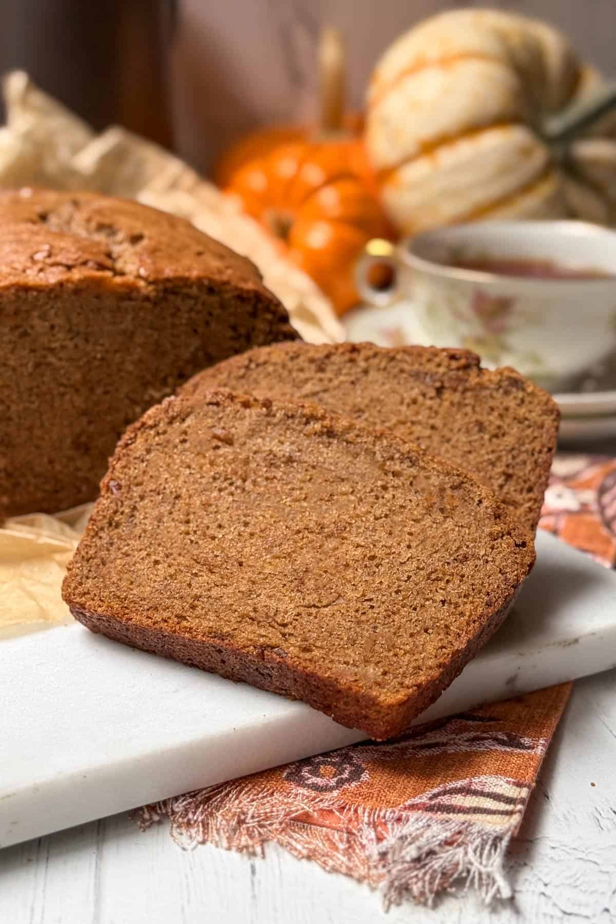Two slices of sweet potato bread rest on a marble board, surrounded by small pumpkins and a cup of tea. The scene has a cozy autumnal feel with warm orange and brown tones.