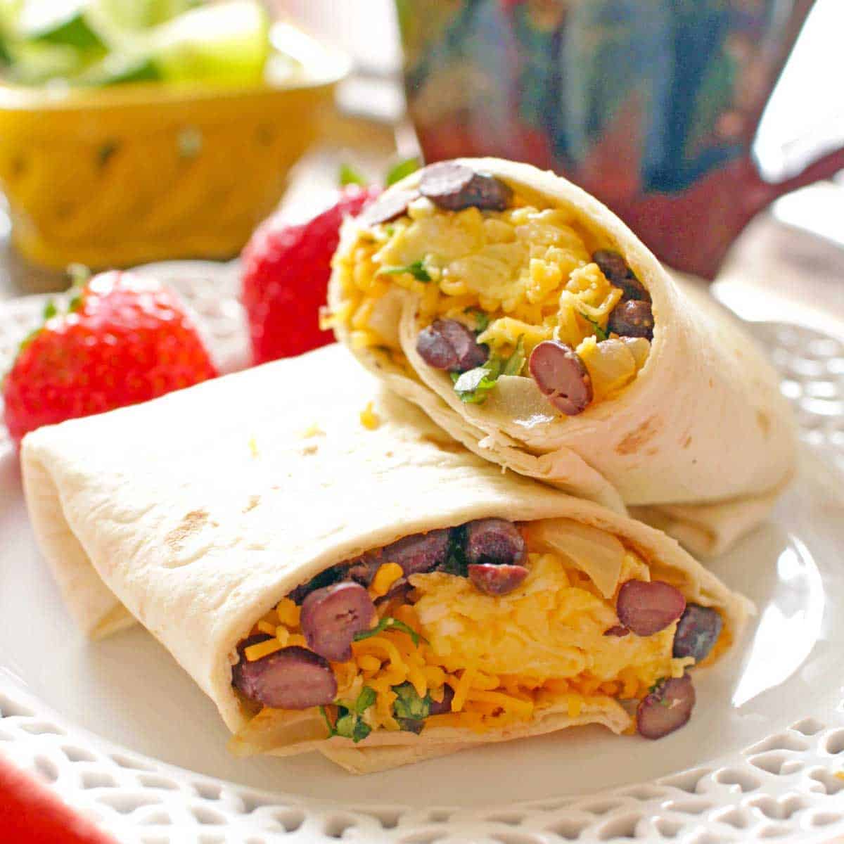 A breakfast burrito cut in half, filled with scrambled eggs, black beans, cheese, and herbs, is placed on a white plate. Fresh strawberries and a blurred background of colorful decor are visible.