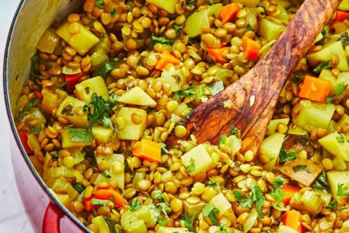 A red pot filled with a colorful lentil and vegetable stew, including carrots, zucchini, and herbs, with a wooden spoon. A small dish with chopped herbs is in the background on a white marble surface.