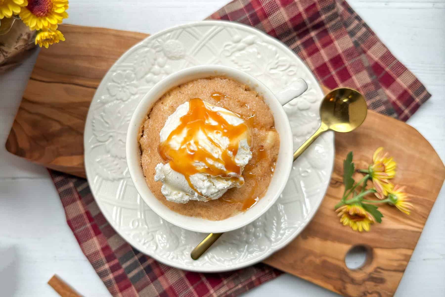 A white mug containing a light brown apple cake topped with a dollop of ice cream and drizzled with caramel sauce. The bowl is placed on a white plate with embossed designs, accompanied by a gold spoon on a wooden board with a red plaid cloth and yellow flowers.