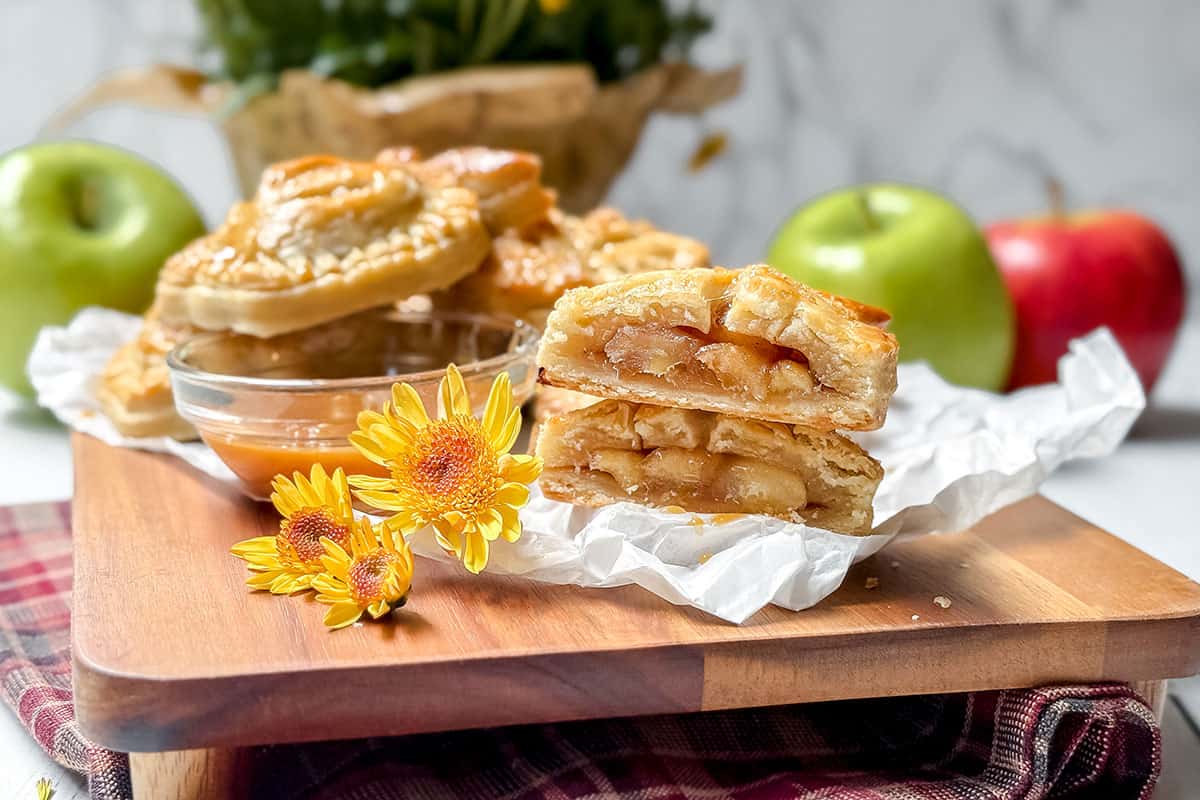 Close-up of apple pie slices on a wooden board, surrounded by fresh green and red apples. The pie is cut to show its filling. Nearby, a small glass bowl with caramel, garnished with yellow flowers, complements the delightful array of apple hand pies on a checkered cloth.