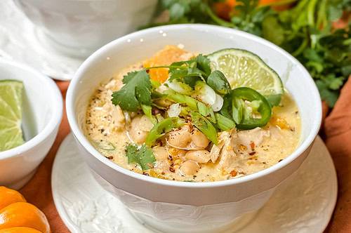 A close-up of a creamy chicken chili served in a white bowl, garnished with lime slices, cilantro, and jalapeño slices. The bowl sits on a white plate with an orange cloth underneath. In the background are small orange pumpkins, cilantro sprigs, and another bowl of the same chili.