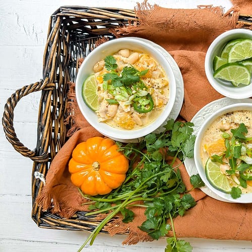 A wicker tray holds two bowls of white chicken chili garnished with lime, cilantro, and jalapeño slices. Alongside, there are lime wedges in a small dish, a bunch of cilantro, mini orange pumpkins, a jar of red pepper flakes, and an orange cloth.