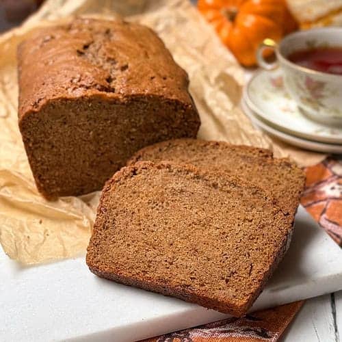 A loaf of sweet potato bread, partially sliced, rests on parchment paper. The background features a teacup filled with red tea, autumn-themed fabrics, and small decorative pumpkins, creating a cozy fall atmosphere on a white table.