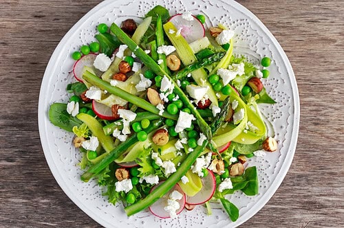 A colorful salad on a white plate featuring asparagus, radishes, peas, mixed greens, hazelnuts, and crumbled goat cheese.