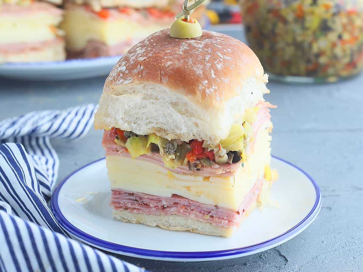A stacked sandwich on a plate with layers of sliced meats, cheese, and pickled vegetables, topped with a thick bun and an olive. A jar of pickled vegetables and a striped cloth are in the background.