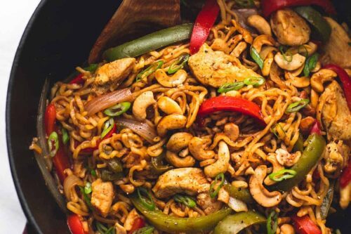 A skillet filled with stir-fried noodles, sliced chicken, bell peppers, cashews, and green onions. A wooden spoon is resting in the skillet, ready to serve.