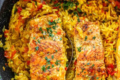 A pan filled with vibrant yellow rice mixed with red and green bell peppers, topped with seasoned salmon fillets sprinkled with chopped parsley. A spoon is partially visible under one salmon fillet.